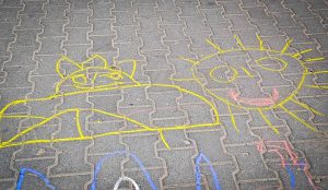 Na kostce brukowej znajdują się rysunki wykonane kredą. Po prawej stronie znajduje się słońce z oczami, nosem i ustami. Po lewej postać.