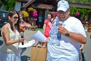 Na pierwszym planie po prawej stoi mężczyzna, w lewej ręce trzyma loda w prawej ma papierową torbę i kartkę. Po lewej stronie profilem stoi kobieta, w prawej ręce ma mikrofon, a w lewej kartki. W tle znajdują się inne osoby oraz budynek z napisem „Zacisze”.