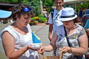 Na pierwszym planie stoją dwie kobiety, pani po prawej trzyma szeroko otwartą papierową torbę. Pani po lewej w prawej ręce trzyma małą żaglówkę. W tle widać inne osoby, drzewa oraz rośliny w donicach.