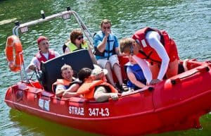 Na pontonie na wodzie znajduje się osiem osób w kapokach. Na samym przodzie stoi mężczyzna reszta osób siedzi.