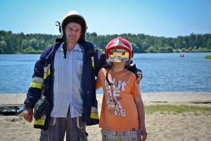 Na pierwszym planie stoją – od prawej: chłopiec w kasku strażackim oraz mężczyzna w kasku i kurtce strażackim. W tle widać piaszczystą plażę, wodę oraz drzewa.