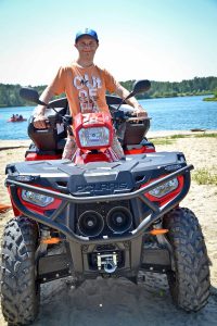 Na quadzie na plaży stoi chłopak, rękami trzyma kierownicę. W tle widać wodę oraz drzewa. Chłopak uśmiecha się szeroko.