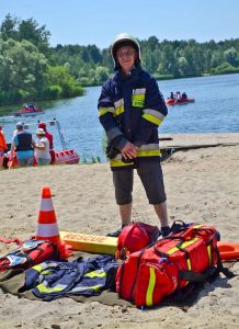 Na plaży stoi osoba ubrana w kask i kurtkę strażacką. Przed nią na kocu rozłożony jest sprzęt ratowniczy. W tle znajduje się jezioro – na nim dwa rowerki wodne a dalej drzewa. Po lewej w tle stoi grupa osób a obok nich znajduje się ponton.