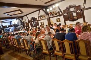 Duża grupa osób siedzi po obu stronach długiego stołu. Na środku zdjęcia tyłem do obiektywu, przy stole, siedzi osoba poruszająca się na wózku. W tle znajduje się ściana a na niej dwa lustra w kształcie okien z okiennicami.