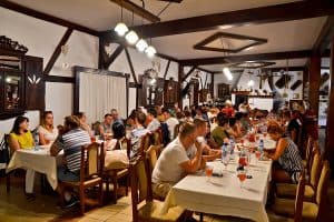 Po obu stronach dwóch długich stołów siedzi duża grupa osób. Stoły przykryte są jasnym obrusem, a na nim znajdują się napoje w dzbankach i szklankach.