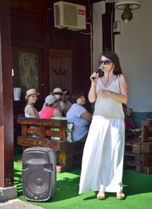 Na pierwszym planie przy głośniku stoi kobieta, w prawej ręce ma mikrofon – jest on skierowany w kierunku jej ust. Za nią tyłem na ławkach siedzi grupa osób – niektóre odwracają głowy w jej kierunku.