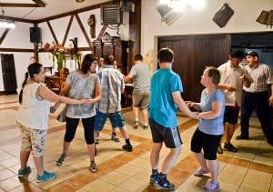 Grupa osób tańczy parami – mniej więcej 10 osób. W tle znajdują się białe ściany oraz duże drewniane drzwi.