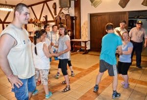 Grupa osób tańczy – mniej więcej 10 osób. W tle znajdują się białe ściany oraz duże drewniane drzwi. Na pierwszym planie po lewej profilem do obiektywu stoi mężczyzna, kciuk prawej ręki trzyma w kieszeni.