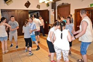 Grupa osób tańczy – mniej więcej 12 osób. W tle znajdują się białe ściany oraz duże drewniane drzwi. Na pierwszym planie cztery osoby stoją w kółku trzymają się za ręce.