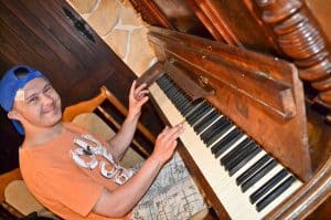 Chłopak siedzi profilem, ale twarz ma skierowaną w obiektyw. Trzyma ręce na klawiszach pianina, które zajmuje prawy kadr – jest ono drewniane. W tle znajdują się duże drewniane drzwi i kamienna ściana.