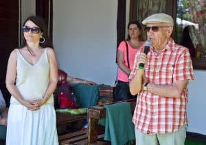 Na pierwszym planie od lewej strony stoją kobieta oraz mężczyzna – trzyma on w prawej ręce mikrofon. W tle znajduje się inna kobieta oraz ściana z oknem i ławki ze stolikiem.