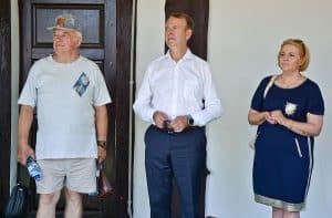 Trzy osoby stoją – są to dwaj mężczyźni oraz kobieta. Pan po lewej w obu rękach trzyma butelki z wodą. W lewej ręce ma jeszcze torbę a w prawej portfel. W tle znajduje się biała ściana oraz drewniane drzwi.