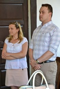 Dwie osoby stoją. Po prawej stronie stoi mężczyzna a po lewej kobieta, trzyma ona założone ręce na brzuchu. W tle znajduje się biała ściana oraz duże drzwi.