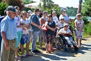 Grupa osób stoi na tle jeziora, białego parasola oraz drzew. Jedna z nich to osoba poruszająca się na wózku. Wszyscy stoją na chodniku.