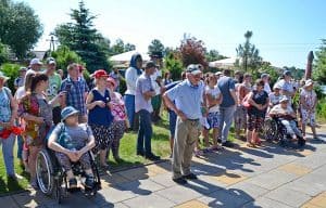 Grupa osób stoi na tle drzew. Dwie z tych osób poruszają się na wózku. Nad nimi widać bezchmurne niebo.