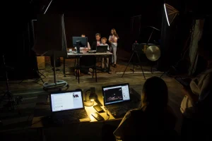 Ciemne pomieszczenie. osoba siedzi przed laptopami, które są włączone. Dalej siedzą ludzie przed monitorami komputerów.