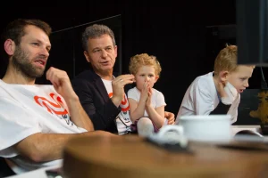 Dwóch mężczyzn siedzi. Obaj mają podniesioną jedną rękę. Obok nich są dzieci.