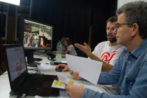 Dwóch mężczyzn siedzi przed biurkiem z komputerami. Jeden z nich trzyma zapisane kartki A4. Drugi ma wyciągniętą rękę i mówi.