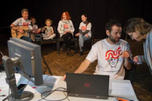 Mężczyzna siedzi przed stołem z komputerami a za nim siedzi grupa osób razem z dziećmi. Jeden z mężczyzn gra na gitarze. Mężczyzna przy komputerach jest odwrócony do kobiety, która pochyla się. Jej sylwetka nie jest cała w kadrze.