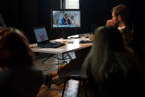 Mężczyzna siedzi przy stole na którym są komputery i patrzy. Na jednym z monitorów jest mężczyzna i dzieci. Dwie kobiety siedzą po prawej i lewej stronie tyłem do obiektywu.