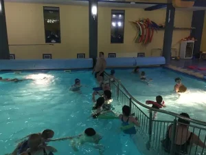 Grupa osób pływa w basenie. Wszyscy są w strojach kąpielowych.