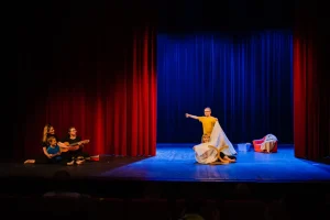 Na scenie przebywają dzieci. Po lewej stronie siedzi dwójka dorosłych z dzieckiem i gitarą.