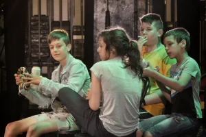 Na scenie przebywa trójka chłopców i jedna dziewczynka siedząca tyłem do obiektywu. Jeden z chłopców trzyma piwo a drugi papierosa.