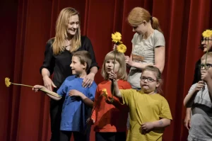 Na scenie stoją dzieci i dwie dorosłe kobiety. Dzieci trzymają żółte kwiaty.