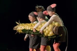 Troje dzieci występuje na scenie. Są przebrane za myszy polne i trzymają długą kolbę kukurydzy.