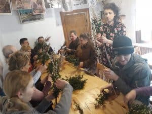 Grupa osób siedzi przy drewnianym stole. Osoby trzymają w rękach gałązki zielone, kwiaty.