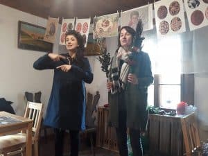 Pośrodku pomieszczenia stoją dwie kobiety. Kobieta po lewej stronie miga, a kobieta po prawej trzyma dwie palmy. Nad nimi wiszą kartki A4, na których są narysowane pisanki.