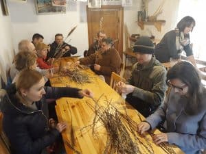 Na stole leżą gałązki bazi. Przy stołach siedzi grupa osób, która sięga po nie. Po prawej stronie stoi kobieta pochylona nad stołem.