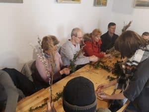 Osoby siedzą przy drewnianym stole. Na nim leżą różne gałązki, a osoby trzymają je w rękach. Na środku siedzi osoba odwrócona tyłem do obiektywu i ma czapkę.