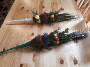 Na stole leżą dwie wykonane palmy. Są zrobione z bazi, kolorowych kwiatów i zielonych gałązek iglastych drzew.