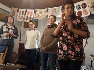 Cztery kobiety stoją na środku pomieszczenia. Kobieta po prawej stronie wykonuje gesty rękoma, miga. Nad kobietami wiszą kartki z namalowanymi pisankami.