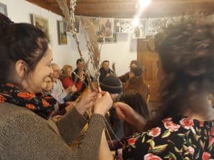 Kobiety trzymają w rękach bazie i trawę pampasową suszoną. Przed nimi siedzi grupa osób.