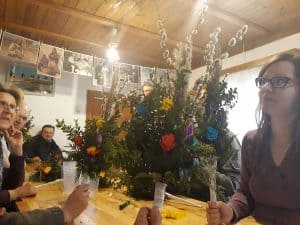 Osoby siedzą przy stole i trzymają gotowe palmy. Każda palma ma bazie, kwiaty oraz białą rączke do trzymania.