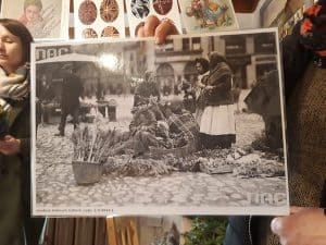 Osoba trzyma w ręce czarno-białe, stare zdjęcie. Są na nim kobiety, a wokół nich są rozmieszczone palmy. Powyżej zdjęcia wiszą kolorowe pisanki narysowane na kartkach.