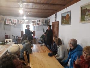 W pomieszczeniu znajduje się grupa osób siedząca przy drewnianym stole po obu stronach. Na krańcu stołu stoją dwie kobiety, a jedna z nich trzyma palmę. Powyżej wiszą narysowane na kartkach pisanki.