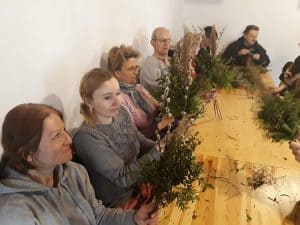 Przy drewnianym stole siedzą osoby, które trzymają zielone gałązki, bazie. Kobieta po lewej stronie uśmiecha się patrząc na gałązki.