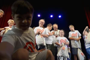 Grupa dzieci stoi na scenie z workami. Wśród nich są także dorośli. Jeden z chłopców jest bardzo blisko obiektywu.