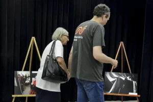 Starsza kobieta i mężczyzna idą razem. Za nimi sztalugi ze zdjęciami.