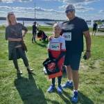 Mężczyzna z małą dziewczynką w strojach sportowych z przypiętymi numerami startowymi pozują do zdjęcia. Obok nich stoi kibicująca im z uśmiechem kobieta.