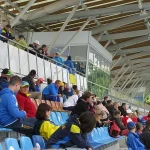 Zawodnicy oraz kibice siedzą na trybunach. Jest ich kilkanaście i mają różnokolorowe kurtki.