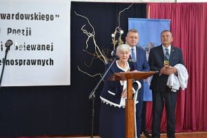 Po prawej stronie stoi za pulpitem starsza kobieta. Ma ona przy sobie mikrofon na statywie. Za nią stoi dwóch mężczyzn w garniturach.