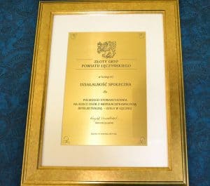 W złotej ramie oprawiona tablica z napisem: “ Złoty Gryf Powiatu Łęczyńskiego w kategorii Działalność Społeczna dla Polskiego Stowarzyszenia na rzecz Osób z Niepełnosprawnością Intelektualną- Koło w Łęcznej. Krzysztof Niewiadomski- Starosta Łęczyński”