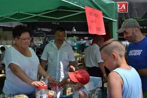 Osoby stoją przed namiotem. Za stołem jest kobieta i mężczyzna a przed nimi stoją dwaj chłopcy i mężczyzna. Na górze jest przyczepiona kartka z napisem “Loteria. Losy”