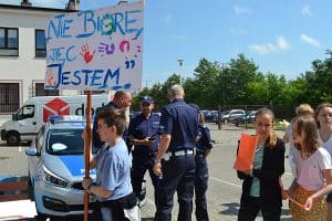 Osoby stoją na zewnątrz. Chłopiec trzyma transparent z napisem:” Nie biorę więc jestem”. Za nim stoją inne osoby i policjanci.