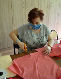 Kobieta w maseczce tnie materiał nożyczkami. Jest on w kolorze różowym.
