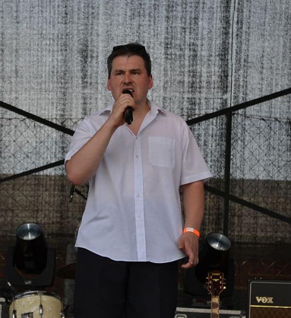 Mężczyzna stoi w białej koszuli i czarnych spodniach na scenie. Mikrofon trzyma blisko twarzy. Za nim stoi perkusja.